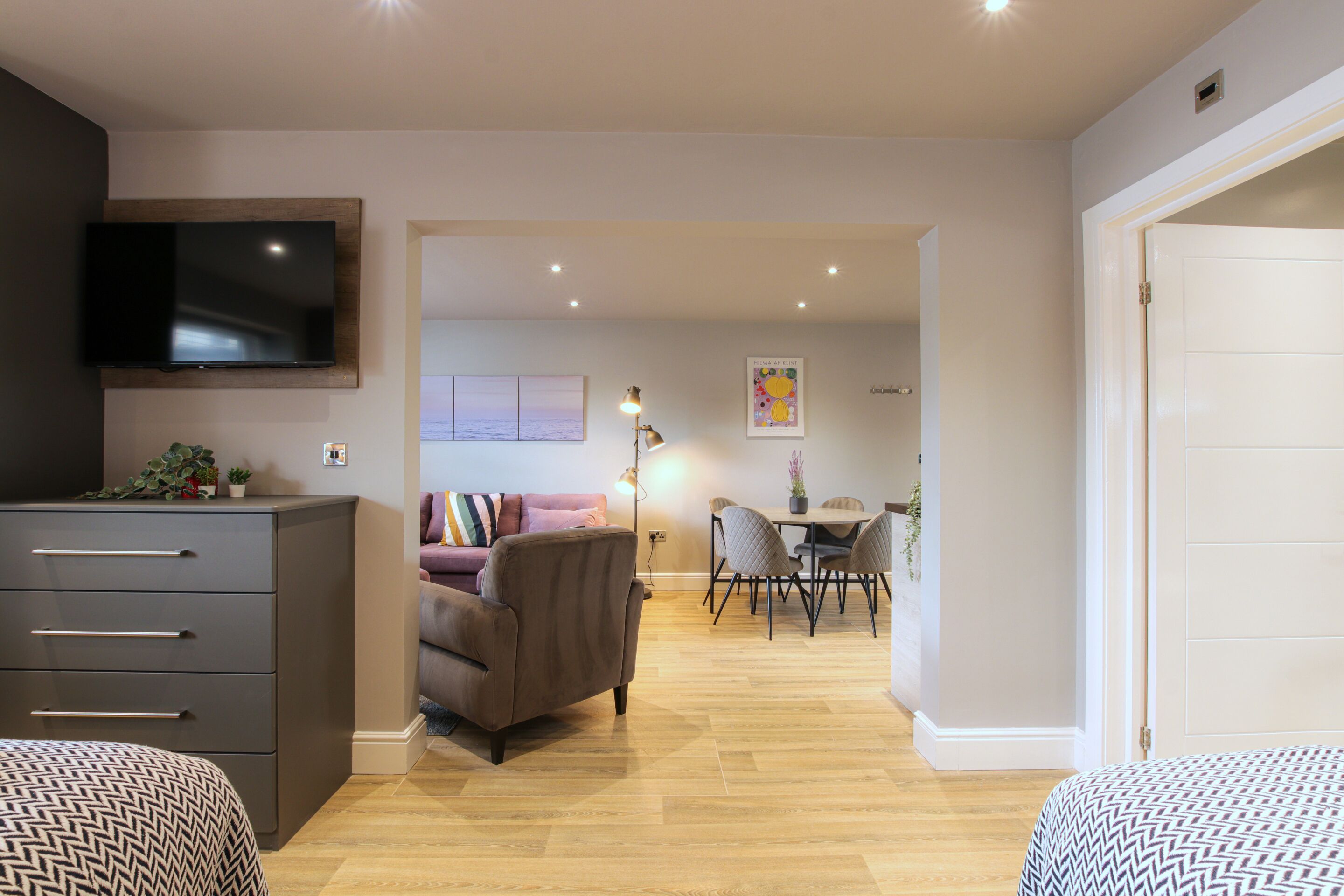 A modern living room with a flat-screen TV, a sofa, a dining area with a table and chairs, and a hallway leading to a bedroom.