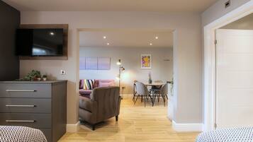 A modern living room with a flat-screen TV, a sofa, a dining area with a table and chairs, and a hallway leading to a bedroom.