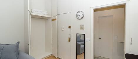 A compact room with a grey sofa, a white wardrobe, a clock on the wall, and a mirror reflecting a grey chair.