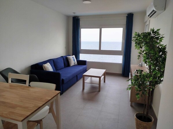 A living room with a sofa, a coffee table, a dining table, and a view of the sea.