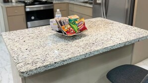 Una cocina con encimera de granito, microondas, estufa y refrigerador. Hay dos taburetes de bar y una cesta de aperitivos en la encimera.