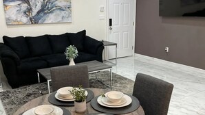 Una sala moderna con un sofá negro, una mesa de comedor redonda con platos y fuentes, una planta pequeña y un cuadro abstracto grande en la pared.