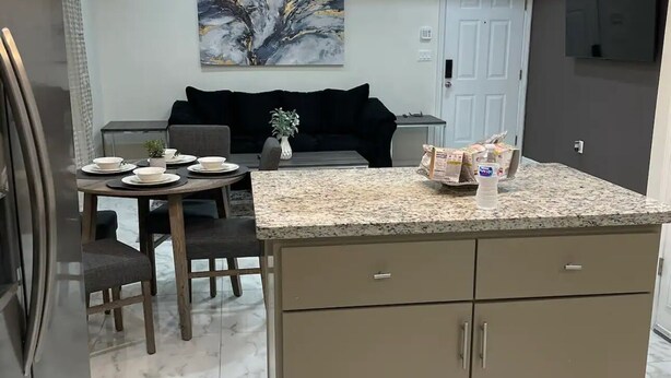 Una cocina moderna con encimera de granito, una zona de comedor con mesa y sillas, y un sofá negro al fondo.