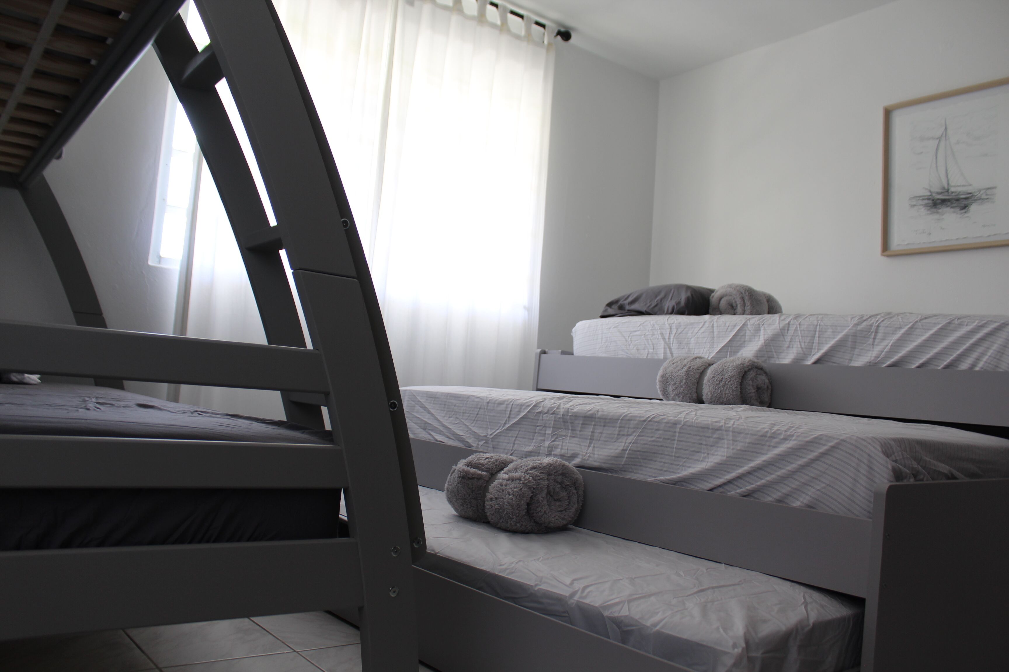Ein Zimmer mit drei Etagenbetten, einem Fenster mit transparenten Vorhängen und einem gerahmten Kunstwerk an der Wand.
