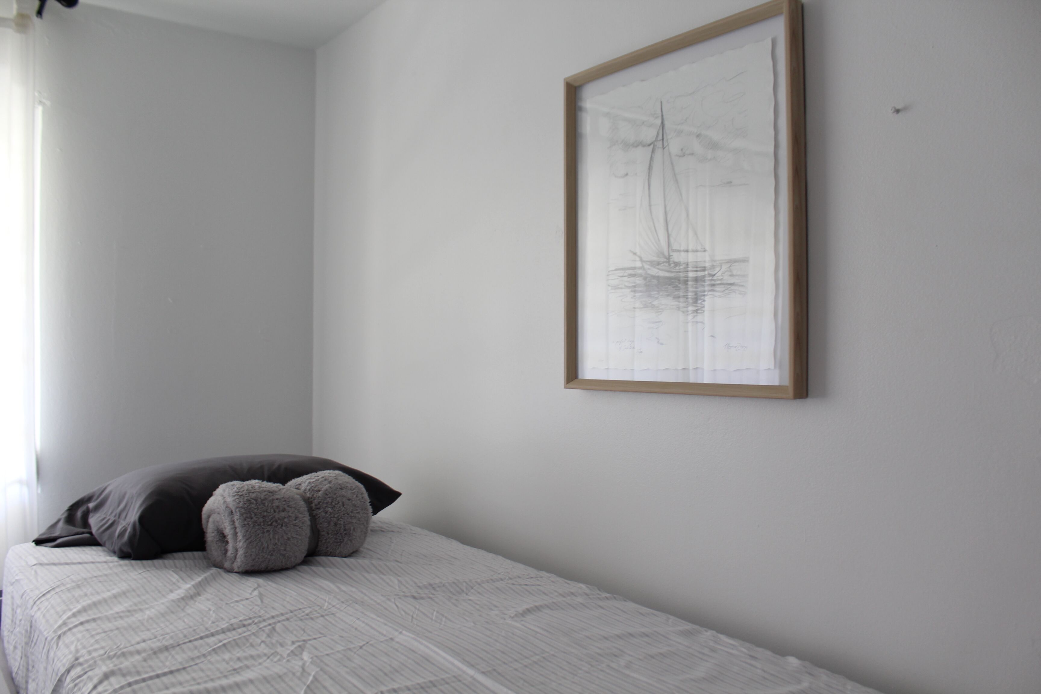 Un dormitorio minimalista con una cama, una obra de arte enmarcada de un velero y una ventana con cortinas.