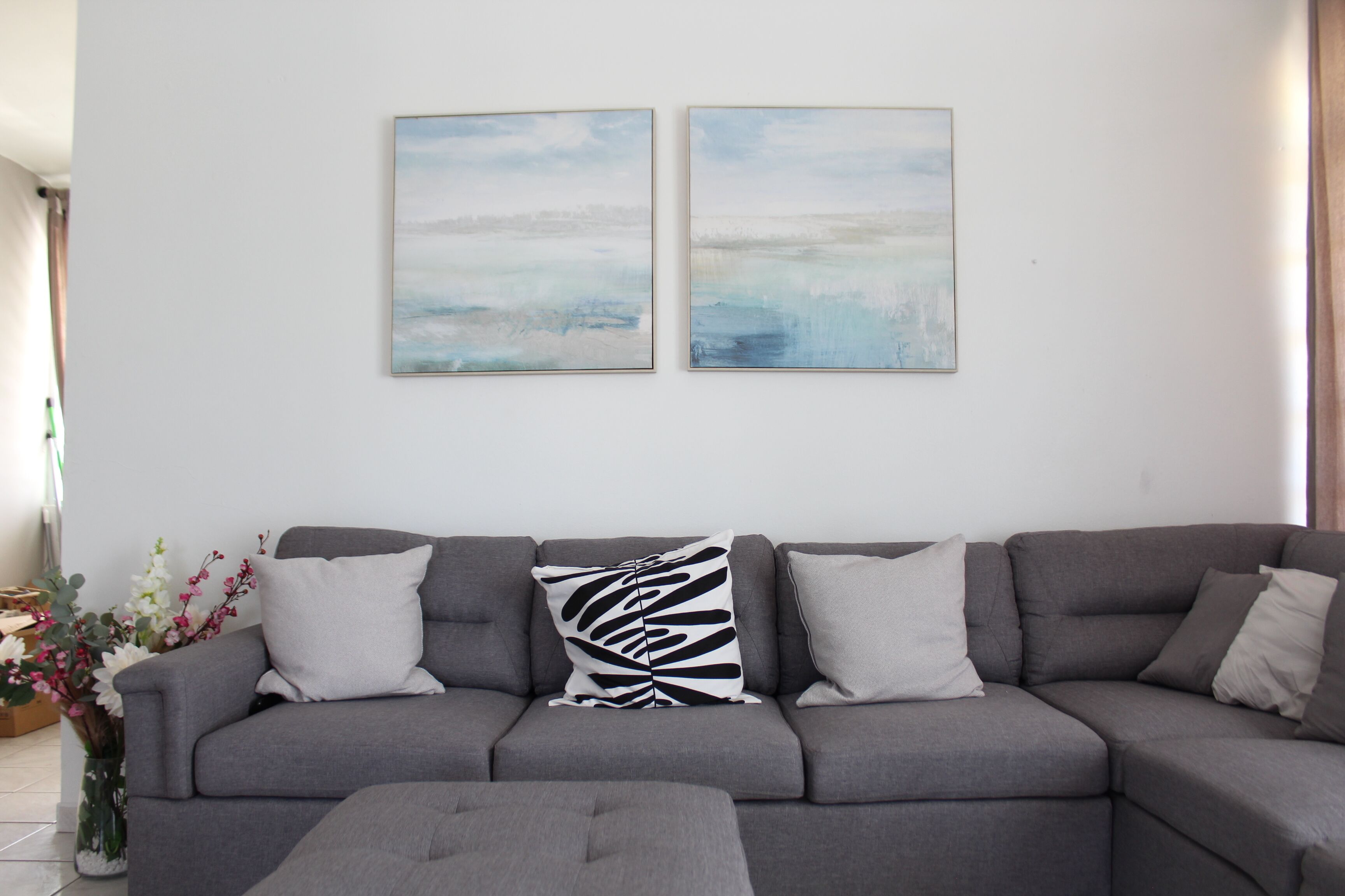 Una sala de estar con un sofá gris, dos cuadros abstractos y un jarrón de flores.