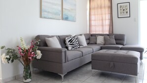 Un salon moderne avec un canapé d’angle gris, un vase en verre contenant des fleurs et une grande fenêtre avec des rideaux.