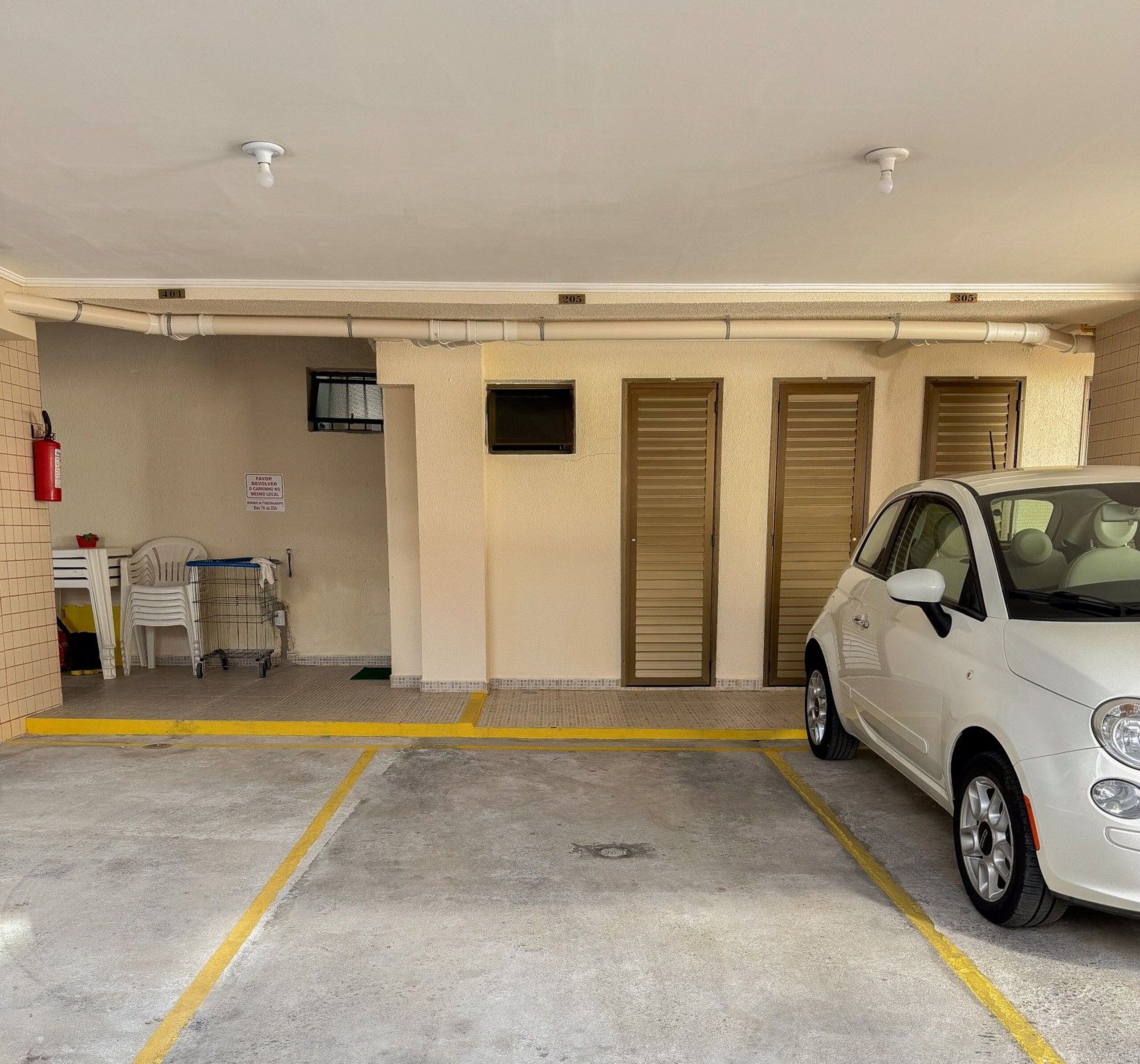 Garagem de estacionamento com um carro branco estacionado numa vaga. Há três portas, um extintor de incêndio e uma placa na parede.