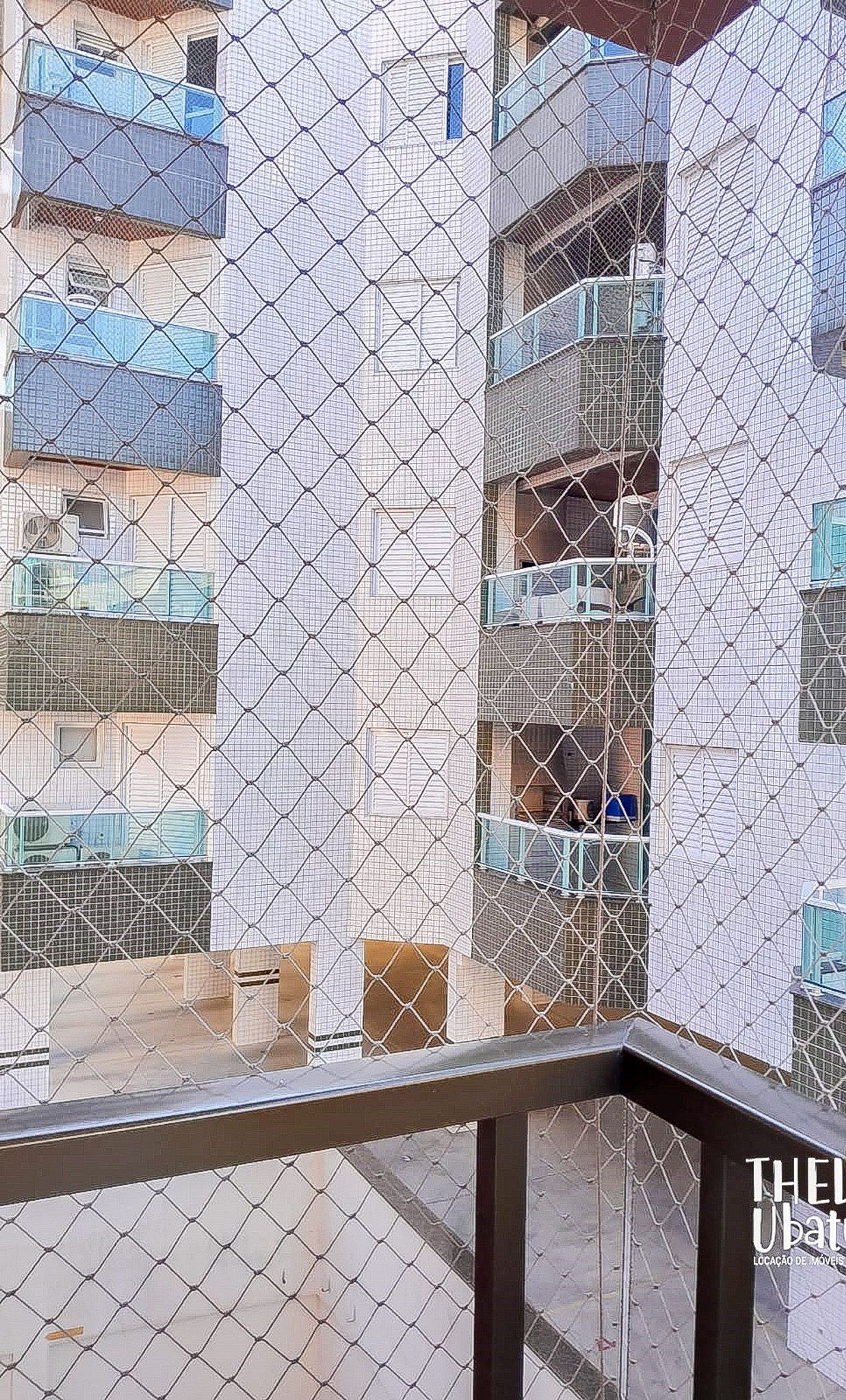 Una valla de malla metálica con barandilla de balcón, con vista a un edificio de varios pisos con balcones.