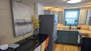 A modern kitchen with a black countertop, a refrigerator, and light blue cabinets. There is a window with blinds and a vase with yellow flowers on the counter. - Lakefront Home with Sunsets, Dock, and Deck!  Dogs Stay Free! (Delton)