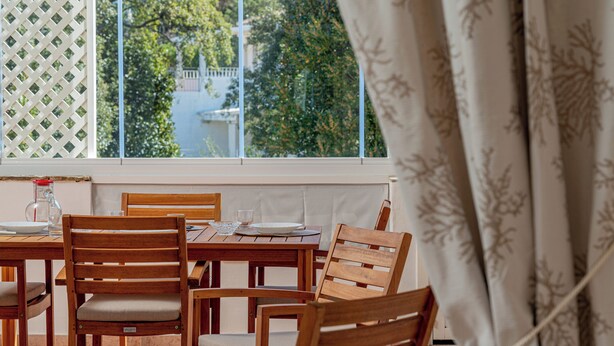 A dining area with wooden chairs and a table, a window with a view of trees and a building, and a curtain partially covering the window. - Sweet Mavy Incantevole Dimora by Wonderful Italy (Olbia)