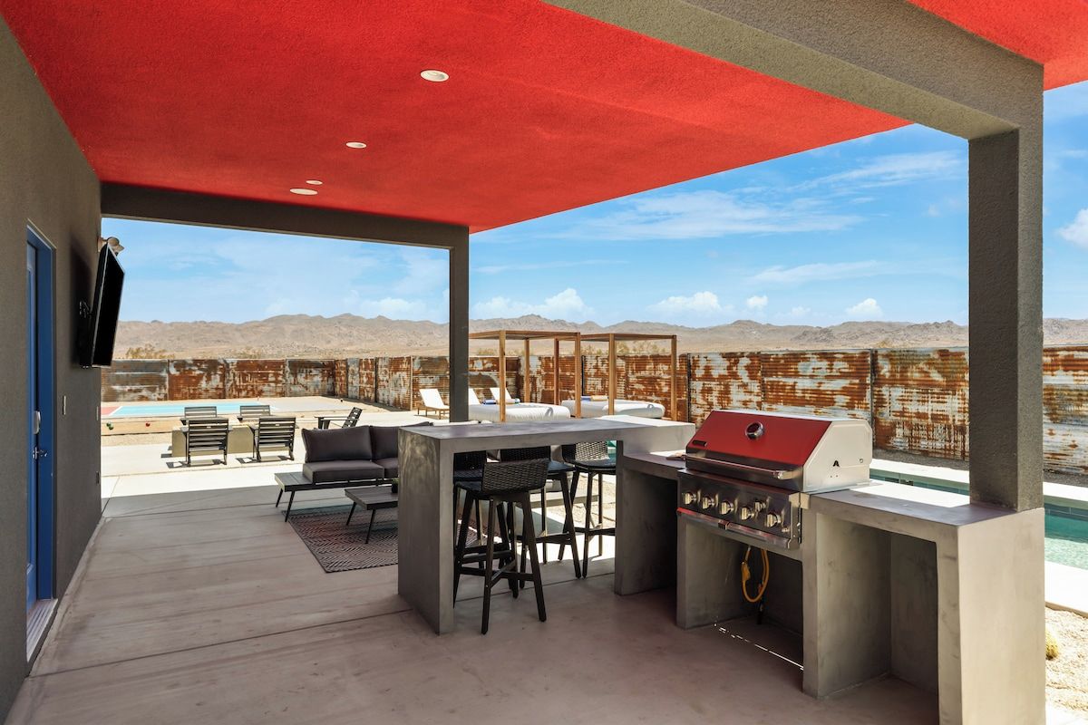 An outdoor patio with a barbecue grill, seating area, and a pool in the background.