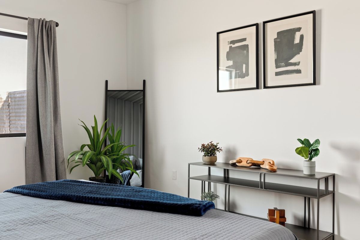 A bedroom with a bed, a bedside table with a plant, a mirror, and two framed artworks on the wall.