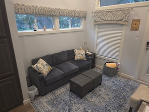 Une pièce chaleureuse avec un canapé gris foncé, un pouf assorti, un tapis à motifs et une fenêtre agrémentée d’une lambrequin décorative.