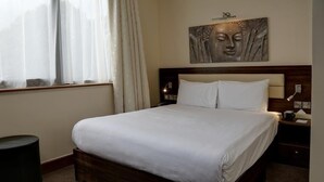 A neatly made bed in a hotel room with a wooden headboard, a bedside table with a lamp, and a wall-mounted artwork.