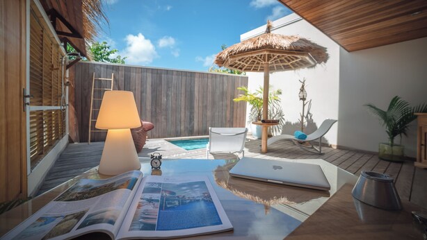 En træterrasse med parasol af strå, en bærbar computer, en bog og et lille bord med en lampe.