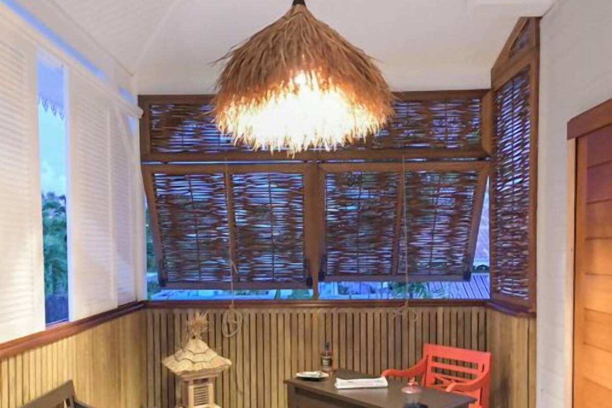 A cozy corner with a wooden bench, a table, and chairs, a hanging light fixture, and a view through a window with bamboo blinds.