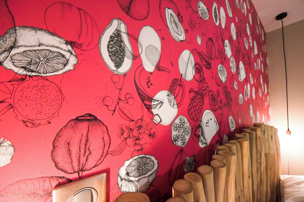 Una habitación con una pared roja vibrante decorada con ilustraciones detalladas de frutas y flores. Se ve un cabecero de madera en primer plano.