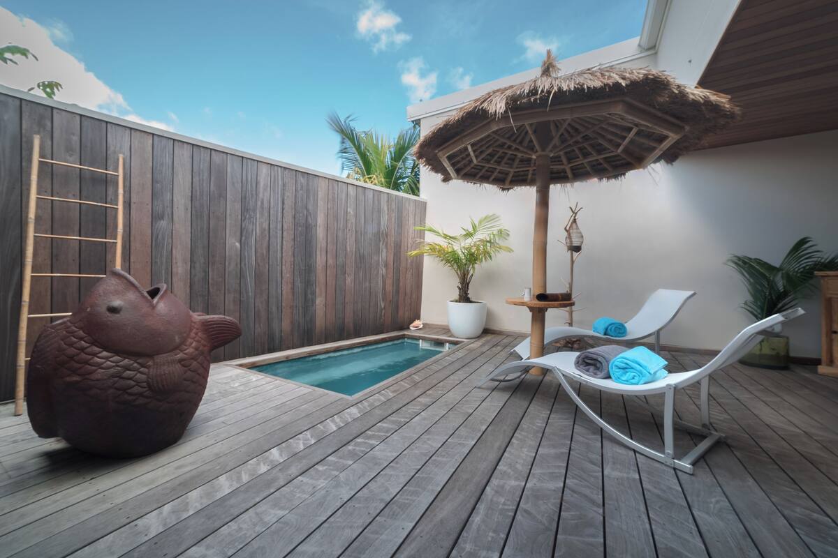 A wooden deck with a pool, lounge chairs, a thatched umbrella, and a decorative fish sculpture.