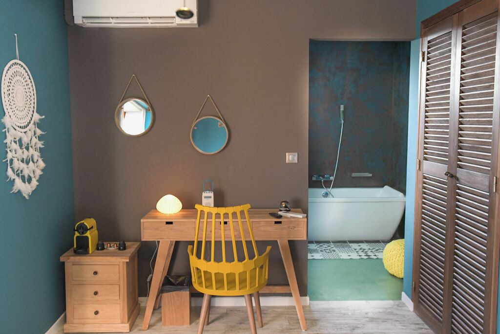 Una habitación con un escritorio de madera, una silla amarilla, una mesita de noche y un espejo adosado a la pared.