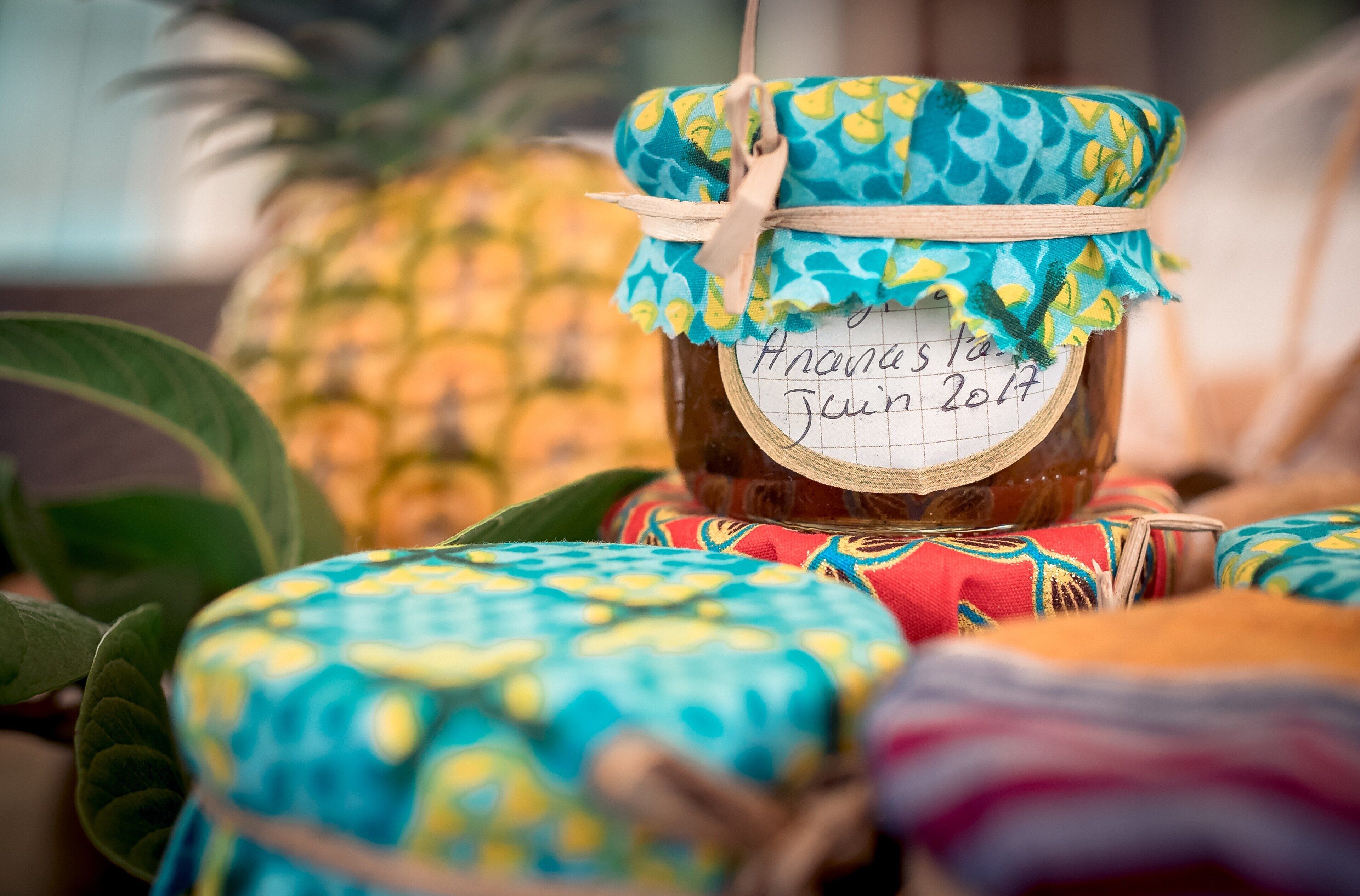 A jar of pineapple jam with a handwritten label, surrounded by colorful fabric items.