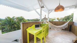 Et lyst, gult bord med en bærbar computer, en hængekøje og en lille pavillon på en træterrasse med udsigt til palmer.