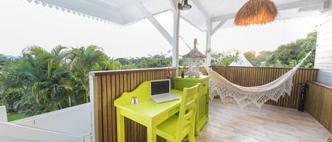Une table jaune vif, un ordinateur portable, un hamac et un petit kiosque sont installés sur une terrasse en bois, d’où l’on peut admirer des palmiers.
