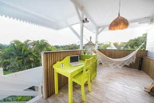 Une table jaune vif, un ordinateur portable, un hamac et un petit kiosque sont installés sur une terrasse en bois, d’où l’on peut admirer des palmiers.