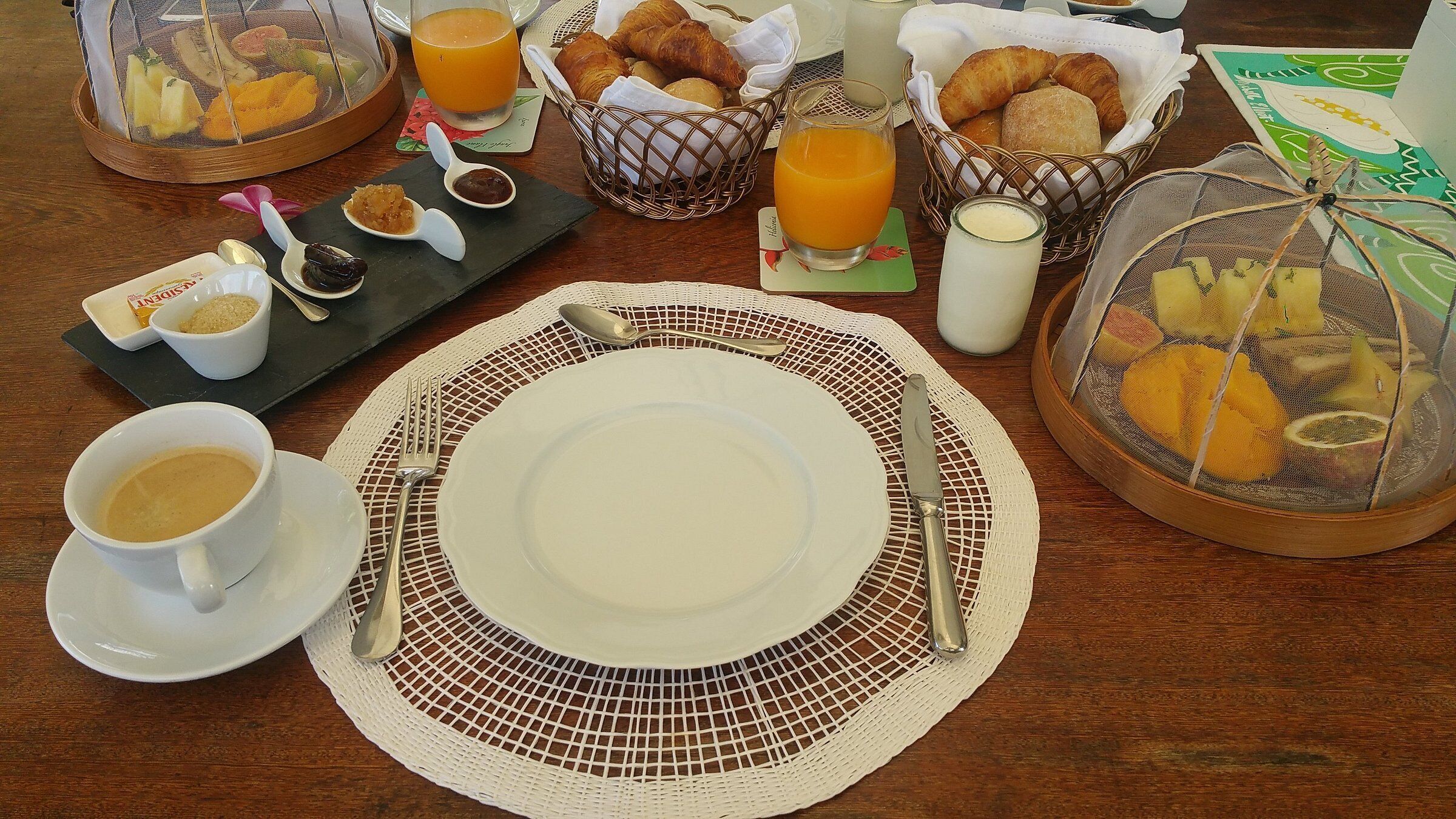 En frukostuppsättning med en tallrik, bestick, en kopp kaffe, croissanter, apelsinjuice och en fruktskål.