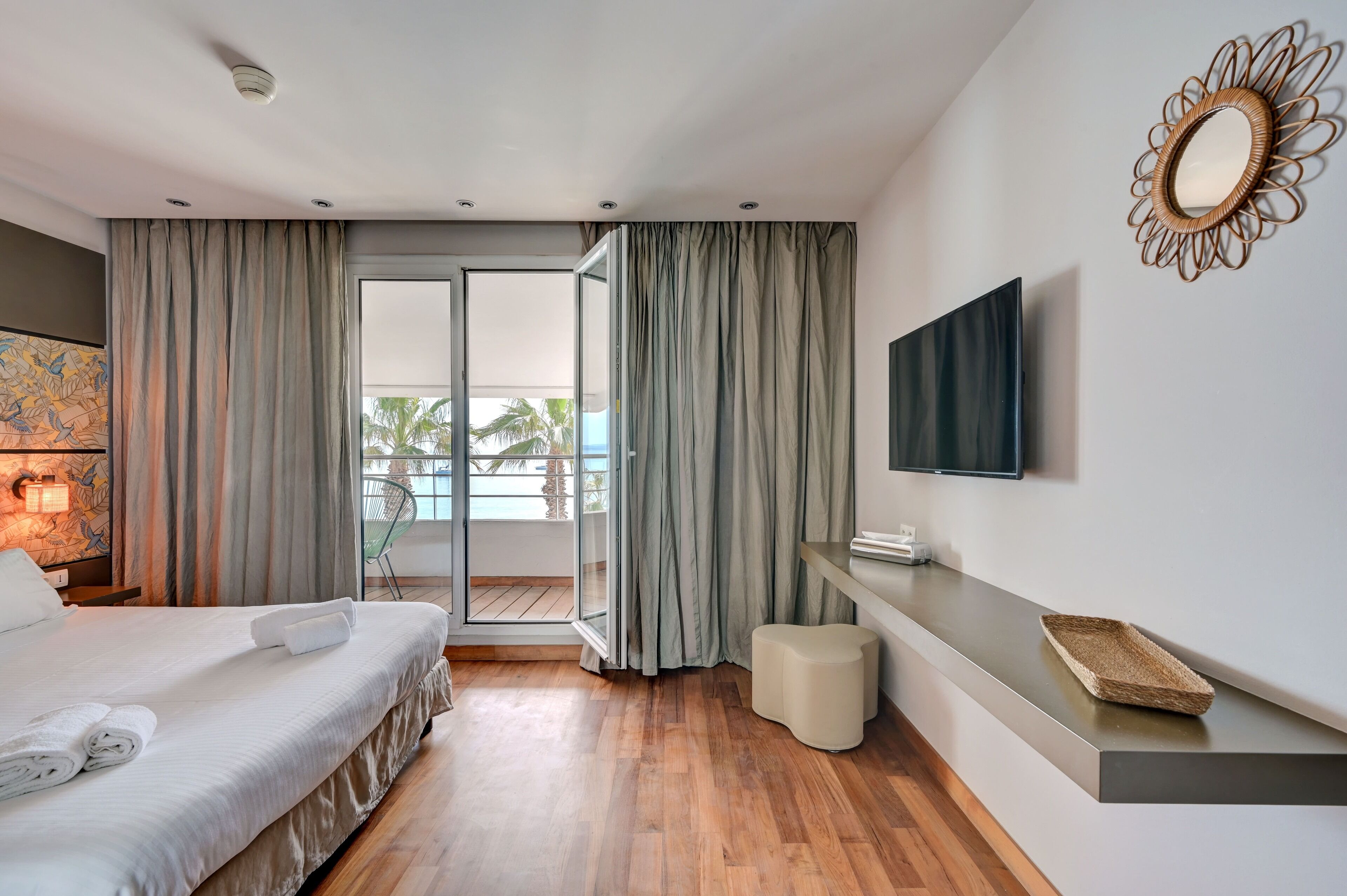 A modern hotel room with a bed, a flat-screen TV, a small table, and a balcony with curtains.