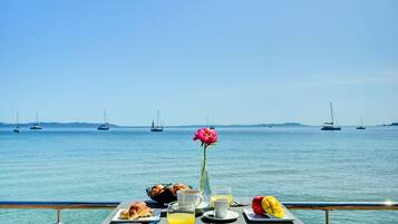 Una mesa con alimentos para el desayuno y una flor, con el mar al fondo y barcos.
