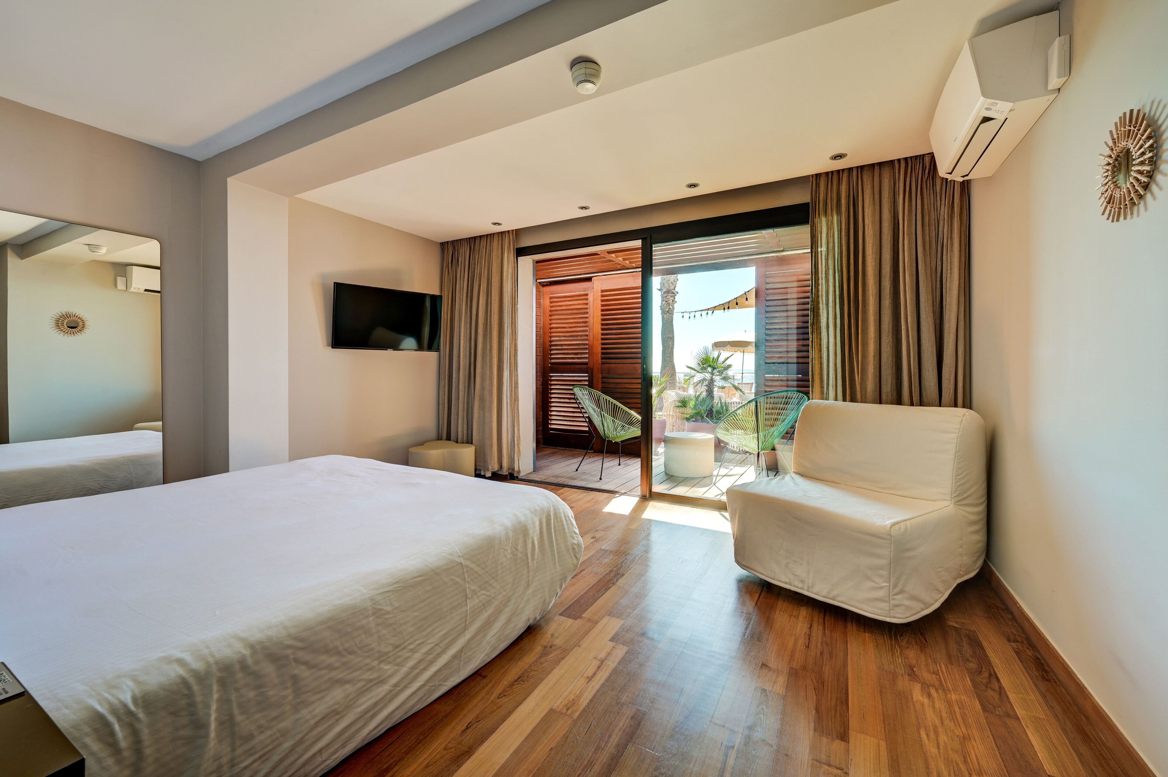 A modern hotel room with a large bed, a seating area with a chair, a TV mounted on the wall, and a sliding glass door leading to an outdoor area.