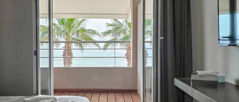 A modern bedroom with a bed, a bedside table, a TV, and a balcony with a view of palm trees and the sea.