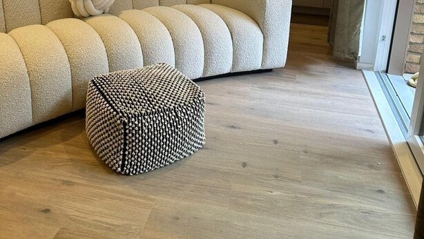 Ein modernes Interieur mit einem Ecksofa und einem dekorativen Hocker, ein Küchenbereich mit Arbeitsplatte sowie ein Essbereich mit Tisch und Stühlen.