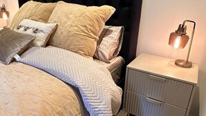 Ein ordentlich bezogenes Bett mit schwarzem Kopfteil, beige farbenen Bettwaren und dekorativen Kissen. Ein Nachttisch mit Lampe und Schublade ist ebenfalls vorhanden.