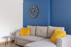 En stue med en beige sofa, to gule puder og et vægmonteret ur.