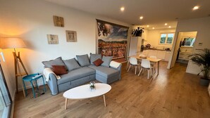 A modern living room with a grey sectional sofa, a small round table, and a dining area with white chairs. - La Pascaline, Appt 47m² 1er Étage Avec Belle vue (Saint-Clément-de-Rivière)