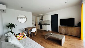 A modern living room with a sofa, coffee table, and a dining area with chairs. There is a kitchen with cabinets and a TV in the living room.