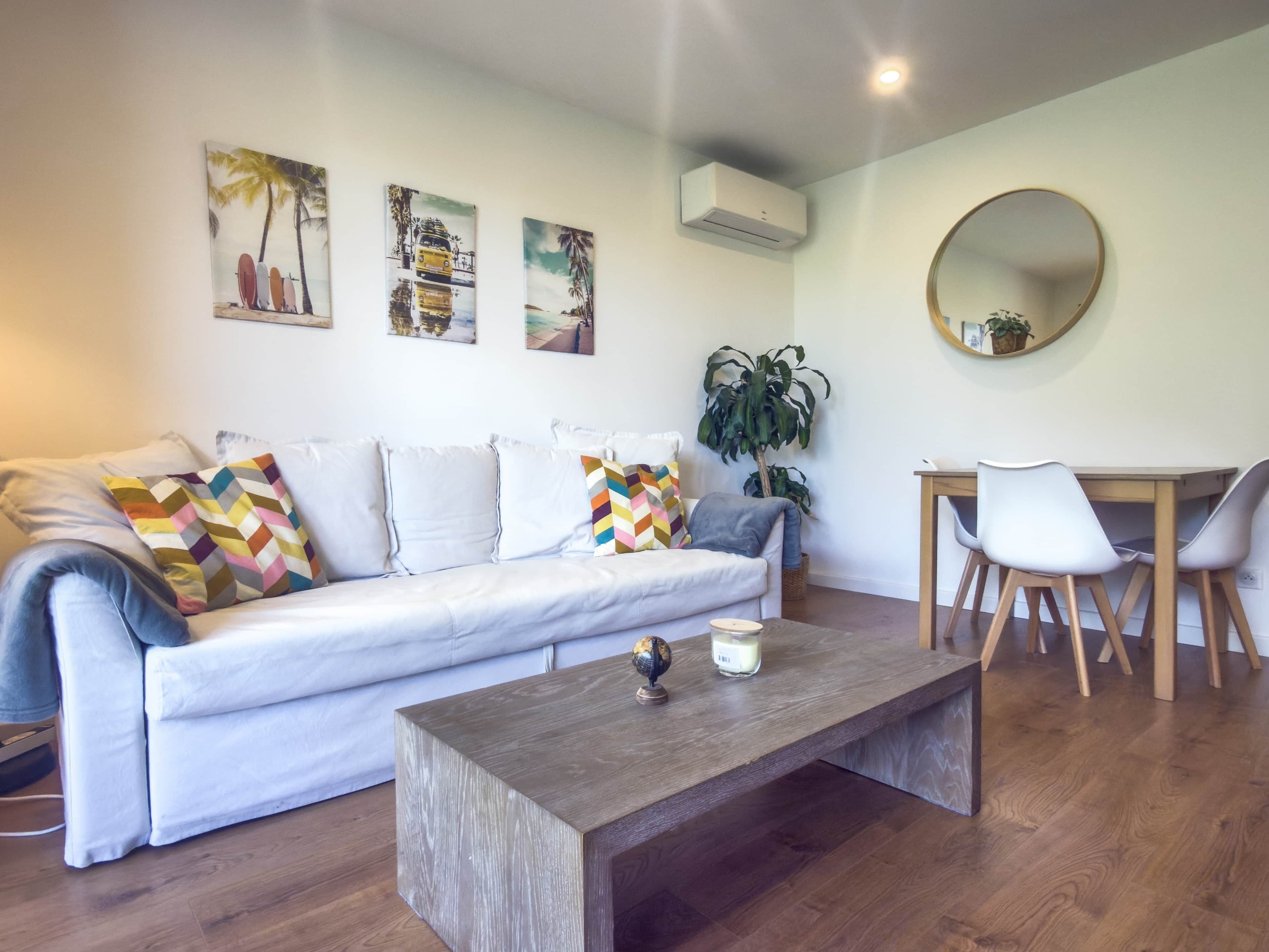 A modern living room with a white sofa, a wooden coffee table, a dining table with chairs, and wall-mounted artwork.