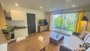 A modern kitchen with white cabinets, a dining table with chairs, a sofa with cushions, and a large window with a view of greenery.