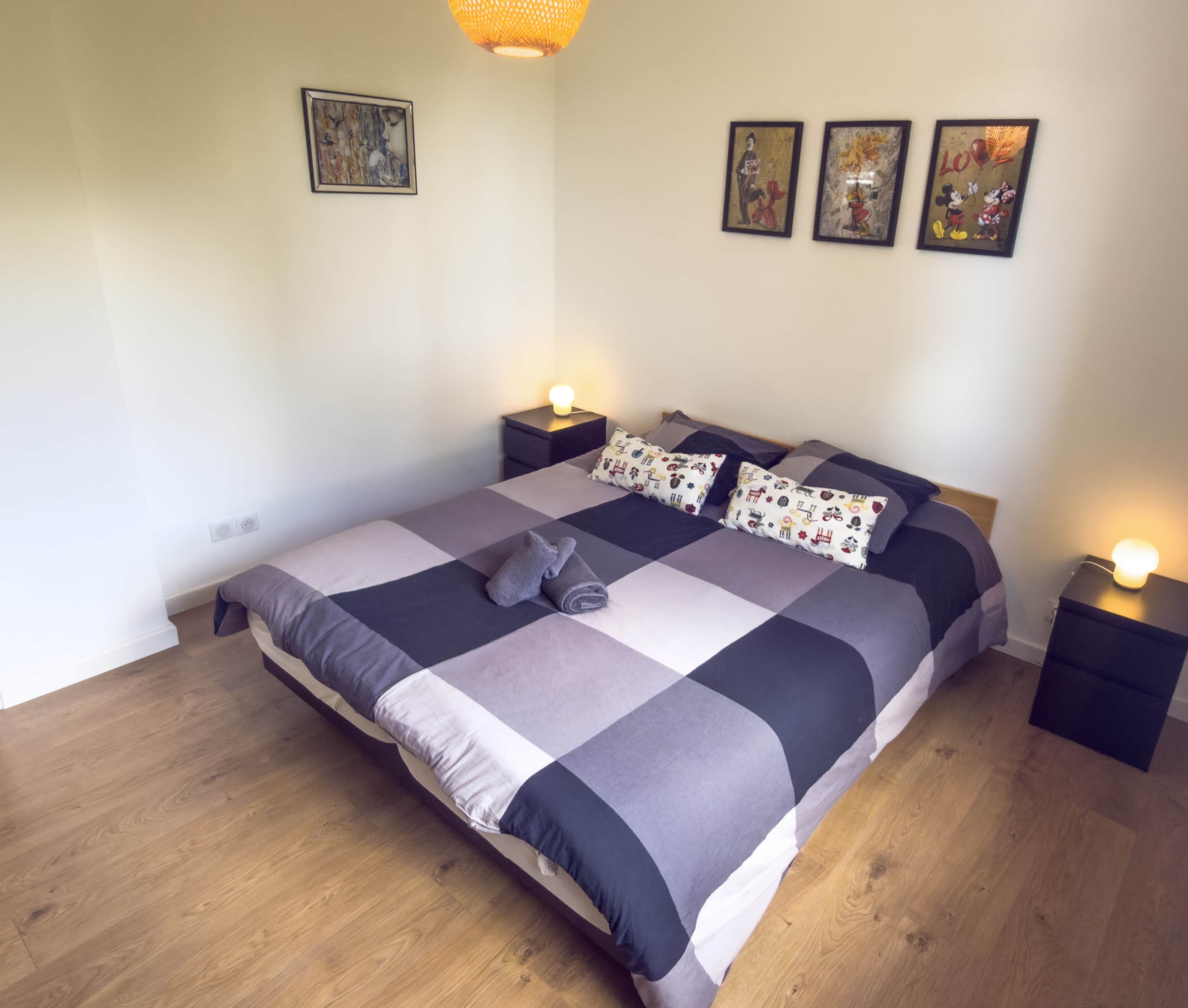 A neatly made bed with a checkered bedspread, two pillows, and a rolled towel on the bed. There are two bedside tables with lamps, and three framed artworks on the wall.