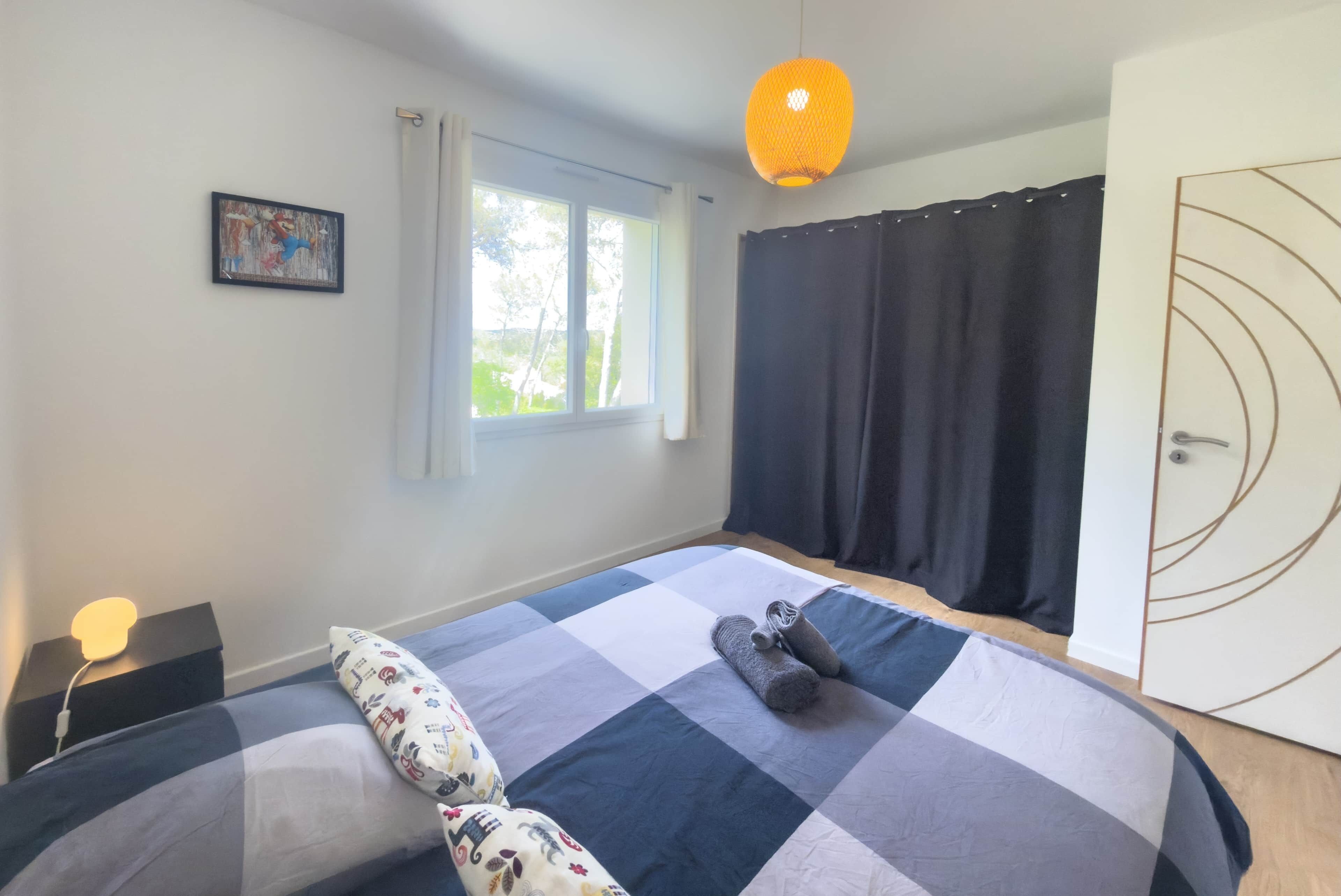 A bedroom with a checkered bedspread, a bedside lamp, a window with curtains, and a hanging light fixture.