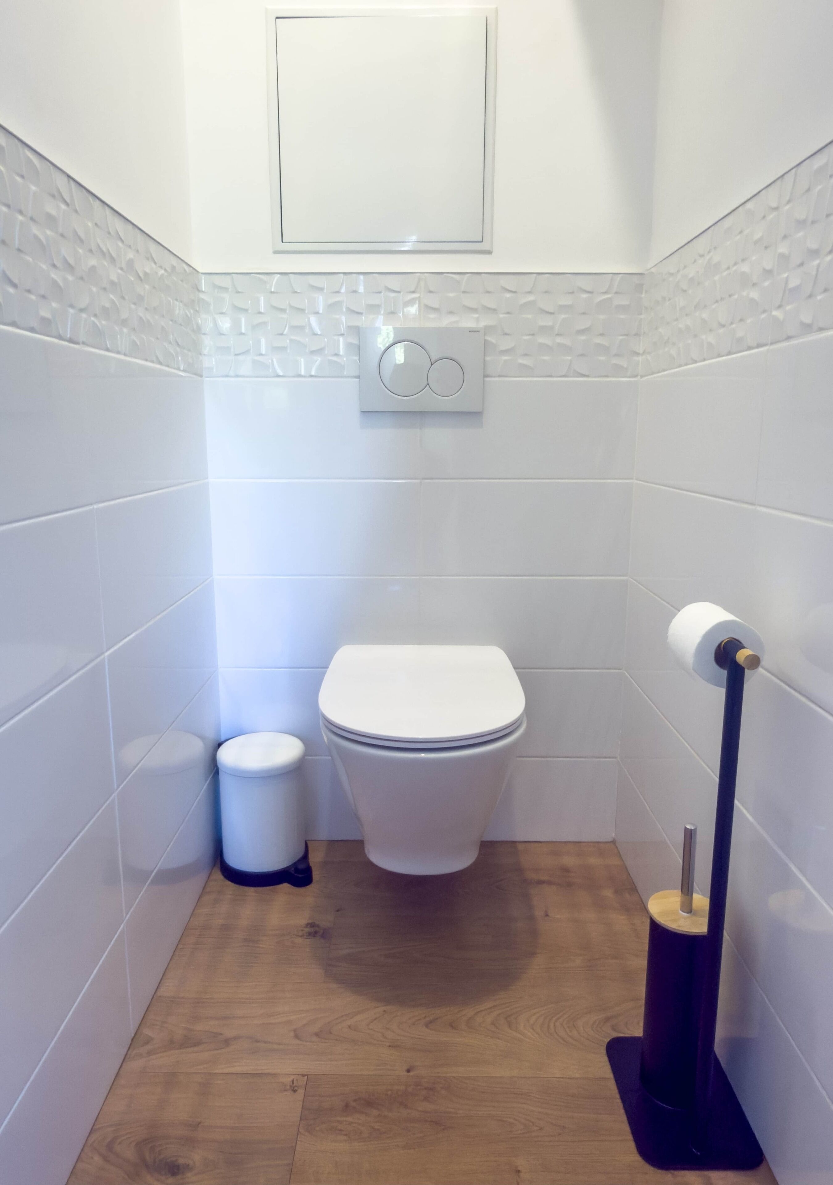 Een kleine, moderne badkamer met witte tegels, een toilet, een prullenbak en een wc-papierhouder.