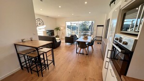 A modern kitchen with a wooden table and bar stools, a dining area with a sofa and chairs, and a large window providing a view of the outdoors. - To complete (Saint-Clément-de-Rivière)