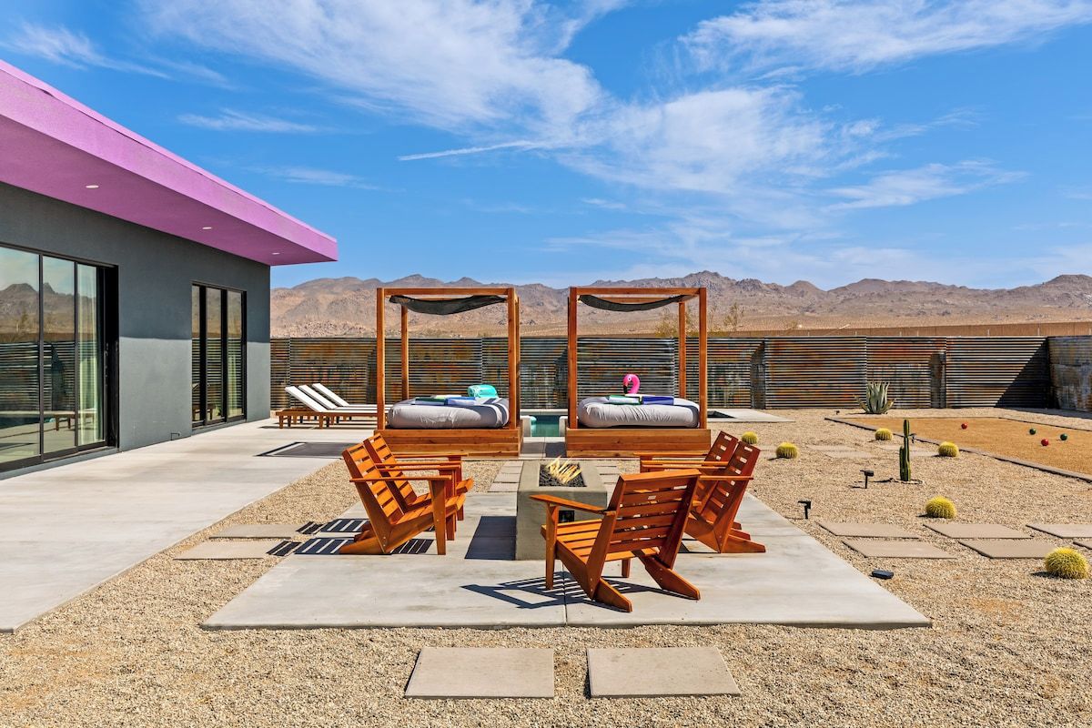 Un área de estar al aire libre con sillas de madera y una mesa, un edificio moderno con techo rosa, y un paisaje desértico al fondo.