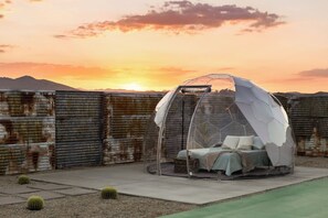 A transparent dome structure with a bed inside, set against a backdrop of a sunset and a rusty metal fence.