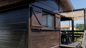 A wooden cabin with a covered porch, a window with shutters, and a view of a house in the background. - Gothes Chalet Javier!! (Ponte de Vagos)