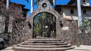 Une entrée en pierre surmontée d’une cloche, des marches menant à cette entrée et une enseigne indiquant « Pueblo Colonial ».