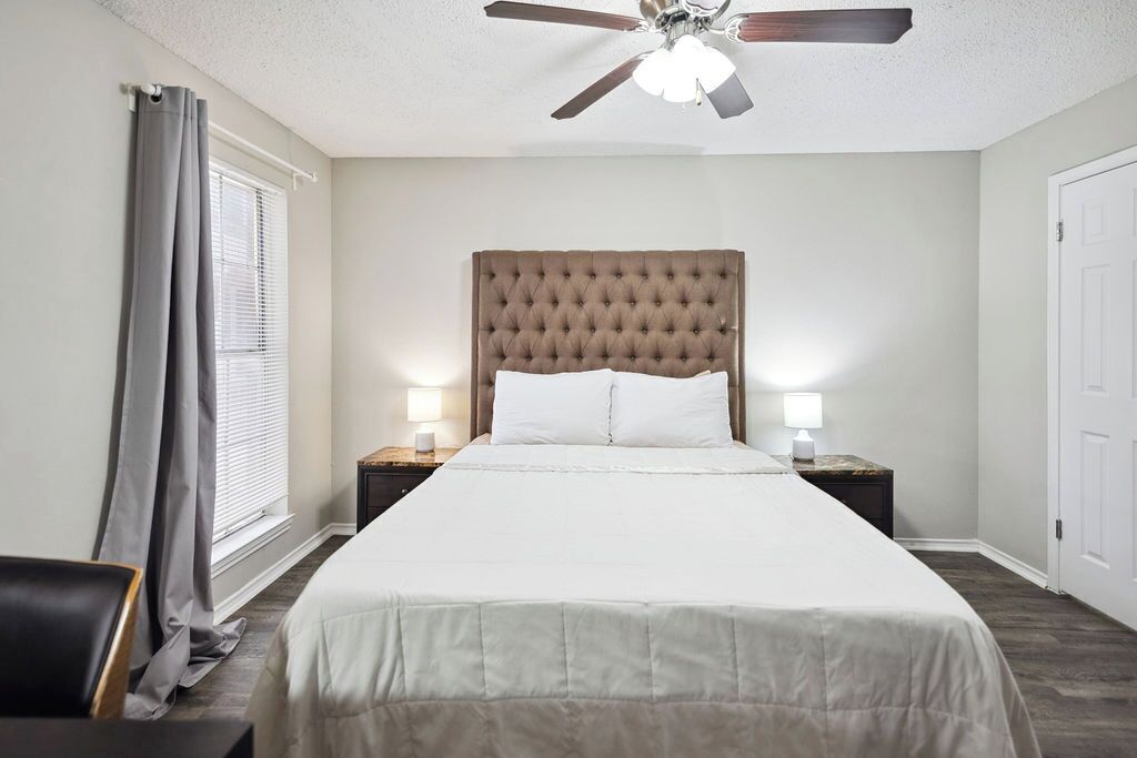 Una habitación de hotel ordenada con una cama grande, dos lámparas de noche, un ventilador de techo y una ventana con cortinas.