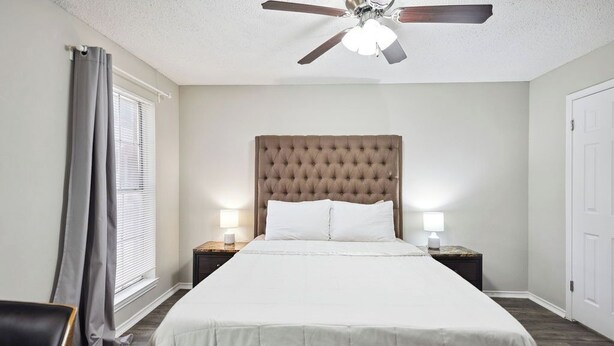 Une chambre d’hôtel bien aménagée, avec un grand lit, deux lampes de chevet, un ventilateur de plafond et une fenêtre avec des rideaux.